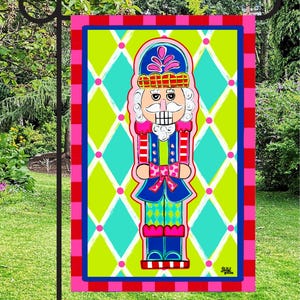 May include: A colorful garden flag featuring a whimsical nutcracker design. The flag has a bright green and turquoise diamond pattern background, a pink and red border, and a detailed illustration of a nutcracker in blue, red, and pink.