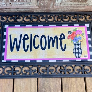 May include: A black welcome mat with a decorative border and a yellow welcome sign with a pink and black checkered border. The sign features a vase of colorful flowers.