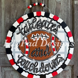 May include: Round wooden sign with a football design. The sign features a brown football with red and white stripes, and the words "Tailgates and Touchdowns" in black script. The border is red, black, and white. A string with red, white, and black beads is attached for hanging.