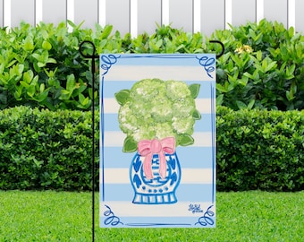 May include: A blue and white striped garden flag featuring a blue and white vase with a pink bow and green hydrangea flowers. The text "Little Garden" is at the bottom of the flag.