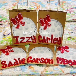 May include: Four brown paper gift bags with white and red painted baseball designs. The bags are decorated with red bows and the names "Izzy Carley", "Sadie", "Carson", and "Megan" are written in red on the bags.