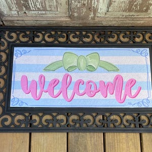 May include: A black welcome mat with a blue and white striped design and a green bow. The word "welcome" is written in pink cursive letters.