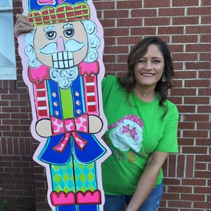 May include: A large, colorful wooden nutcracker decoration. The nutcracker is painted with a pink, blue, green, and red color scheme. It has a detailed face, a hat with a bow, and is held by a person wearing a green shirt and blue jeans.