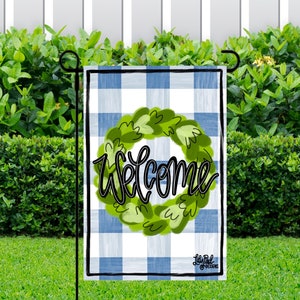 May include: A blue and white checkered garden flag with a green wreath and the word "Welcome" in black script.