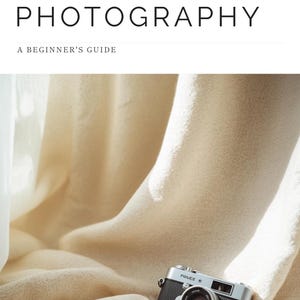 Puede incluir: Una cámara de estilo vintage en negro y plata sobre una tela de color crema suave y drapeada. Sobre la cámara, el texto THE ART OF PHOTOGRAPHY A BEGINNER'S GUIDE aparece en letras mayúsculas negras sin remates sobre un fondo blanco.