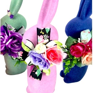 May include: Three Easter bunny figurines, one green, one pink, and one blue, each with a floral arrangement and a ribbon around their necks.