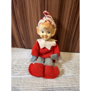 May include: A vintage-style elf doll featuring a red felt outfit, grey felt legs, and red pointed shoes. The doll has a pale face with painted facial features, large ears, and a red and white striped hat with a white trim. The doll is in a seated position.
