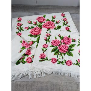 May include: A set of white terry cloth towels featuring a repeating pattern of pink roses with green leaves and stems. The towels have a textured surface and are finished with a fringe along the bottom edges, showcasing a floral design.