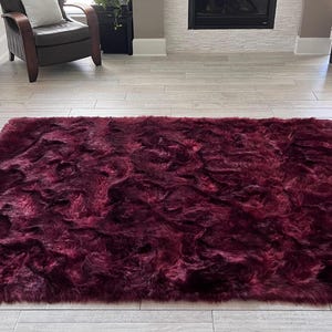 May include: A rectangular, plush burgundy rug with a shaggy texture. The rug is placed on a light-colored wooden floor. The rug is a deep shade of red, with variations in tone, and appears soft and inviting.