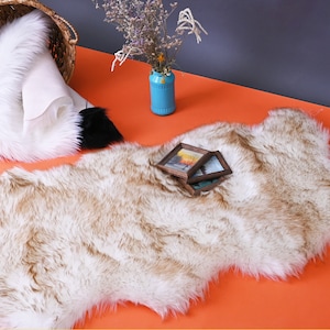 May include: A white and brown faux fur rug with a shaggy texture. The rug is laid out on an orange surface. There are three small picture frames on the rug.
