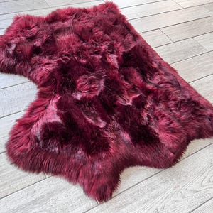 May include: A plush, burgundy-colored sheepskin rug with a natural, irregular shape. The rug has a dense, textured surface with varying shades of red and dark accents, creating a rich, luxurious appearance. The rug is placed on a light-colored wooden floor.