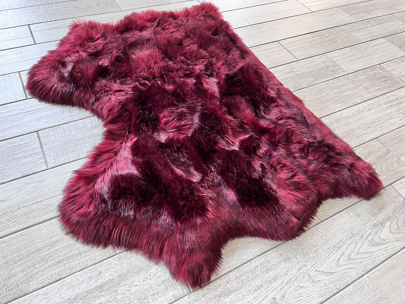 May include: A plush, burgundy-colored sheepskin rug with a natural, irregular shape. The rug has a dense, textured surface with varying shades of red and dark accents, creating a rich, luxurious appearance. The rug is placed on a light-colored wooden floor.