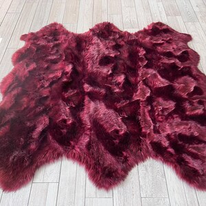 May include: A plush, burgundy-colored faux fur rug. The rug has an irregular shape with a soft, textured surface. The rug is displayed on a light-colored wooden floor.