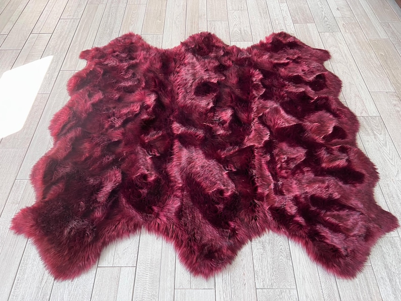 May include: A plush, burgundy-colored faux fur rug. The rug has an irregular shape with a soft, textured surface. The rug is displayed on a light-colored wooden floor.