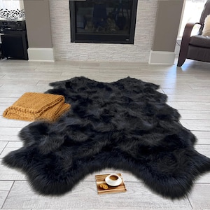 May include: A black faux fur rug with a shaggy texture. The rug is laid out on a light wood floor with a small brown tray and a cup of coffee on top of it. A tan knitted throw blanket is lying on the floor to the left of the rug.