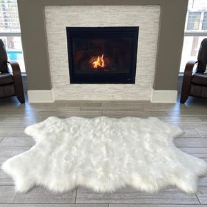 May include: White faux fur rug with a shaggy texture, placed in front of a fireplace in a living room setting.