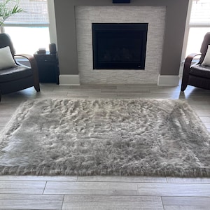 May include: A gray faux fur rug with a long pile, placed in front of a fireplace in a living room with two brown leather armchairs.