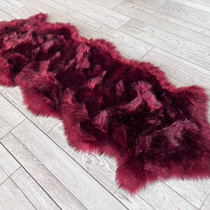 May include: A plush, burgundy-colored faux fur rug. The rug has a long, shaggy pile and an irregular shape. It is placed on a light gray wood-plank floor.