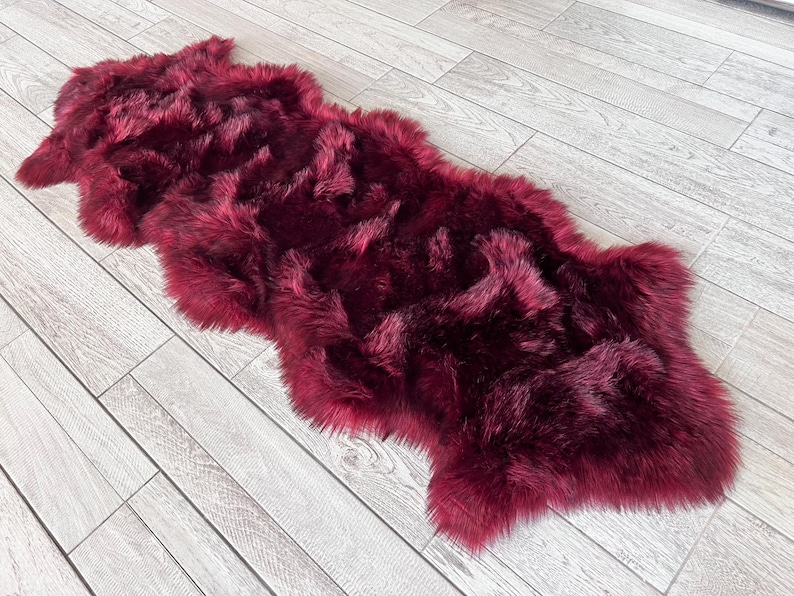 May include: A plush, burgundy-colored faux fur rug. The rug has a long, shaggy pile and an irregular shape. It is placed on a light gray wood-plank floor.