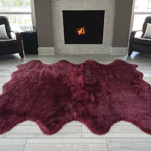 May include: A large, burgundy-colored sheepskin rug with an irregular, organic shape. The rug is placed on a light-colored wooden floor in front of a fireplace and two armchairs.
