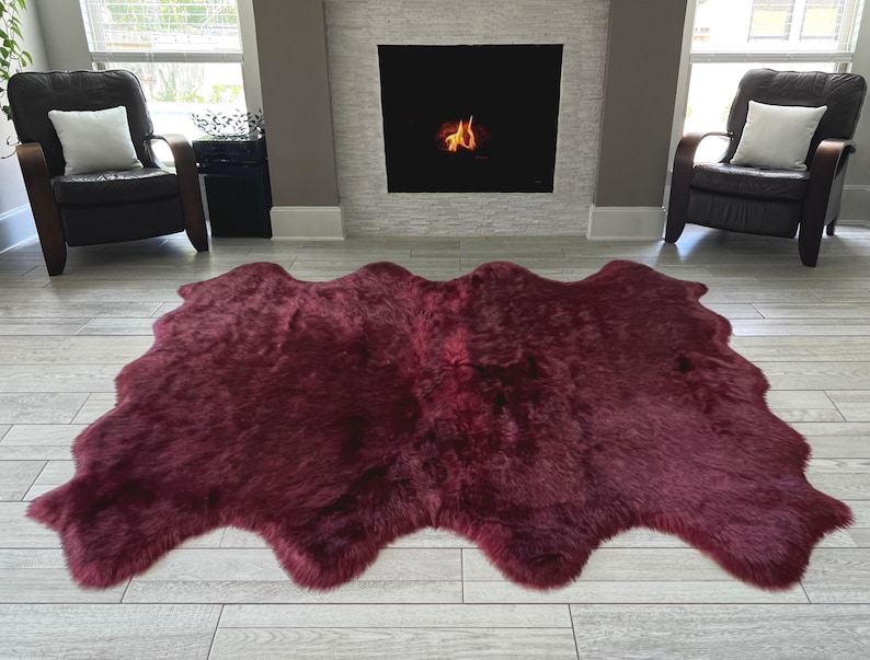 May include: A large, burgundy-colored sheepskin rug with an irregular, organic shape. The rug is placed on a light-colored wooden floor in front of a fireplace and two armchairs.