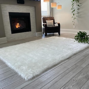 May include: A white faux fur rug is laid out on a wood floor in a living room. The rug is in front of a fireplace with a fire burning inside. There is a chair and a plant in the background.