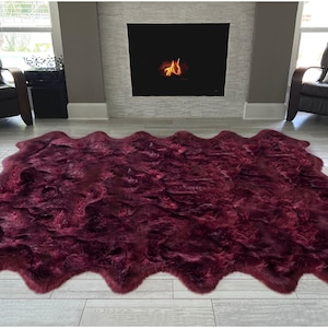 May include: A large, burgundy-colored faux fur rug with a wavy edge. The rug is placed on a light-colored wooden floor in front of a fireplace. Two black leather armchairs with white pillows are on either side.