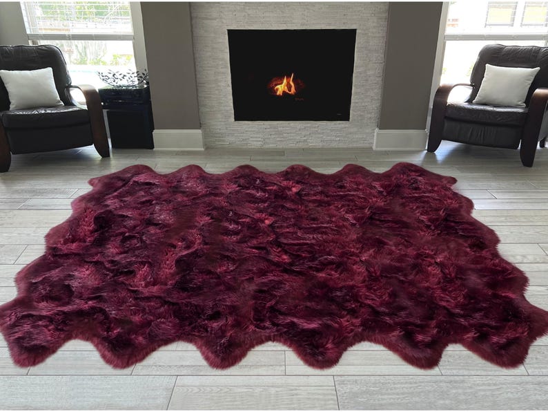 May include: A large, burgundy-colored faux fur rug with a wavy edge. The rug is placed on a light-colored wooden floor in front of a fireplace. Two black leather armchairs with white pillows are on either side.