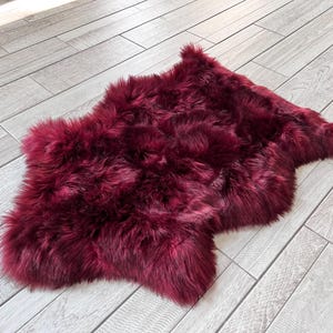 May include: A plush, burgundy-colored sheepskin rug. The rug has a soft, fluffy texture and an irregular shape. The rug is placed on a light gray wooden floor, creating a contrast in color and texture.