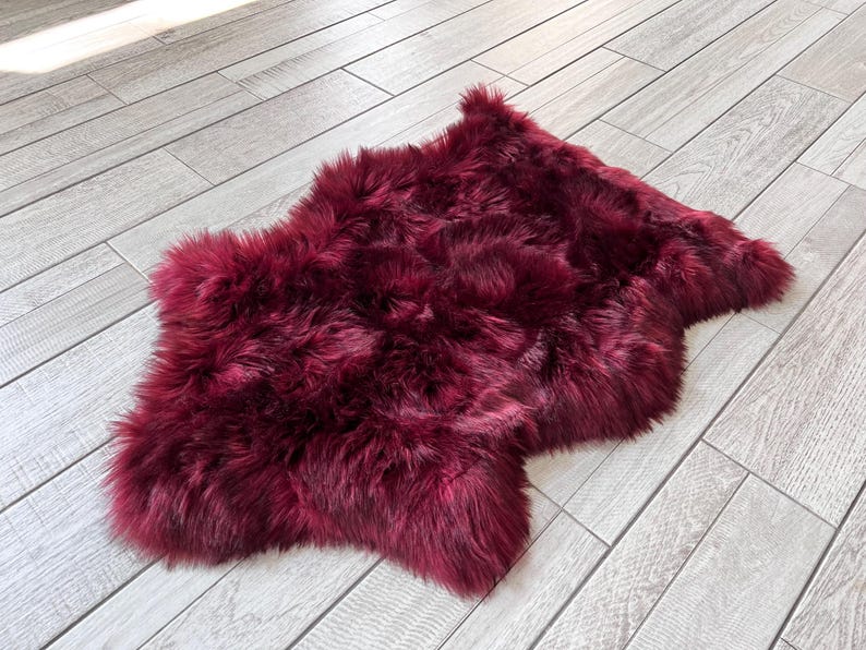 May include: A plush, burgundy-colored sheepskin rug. The rug has a soft, fluffy texture and an irregular shape. The rug is placed on a light gray wooden floor, creating a contrast in color and texture.