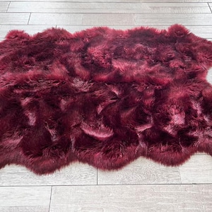 May include: A burgundy-colored sheepskin rug with a soft, textured appearance. The rug is irregularly shaped and lies on a light-colored wooden floor. A small wooden tray with a cup and cookies sits to the left, and a potted plant with pink flowers is on the right.
