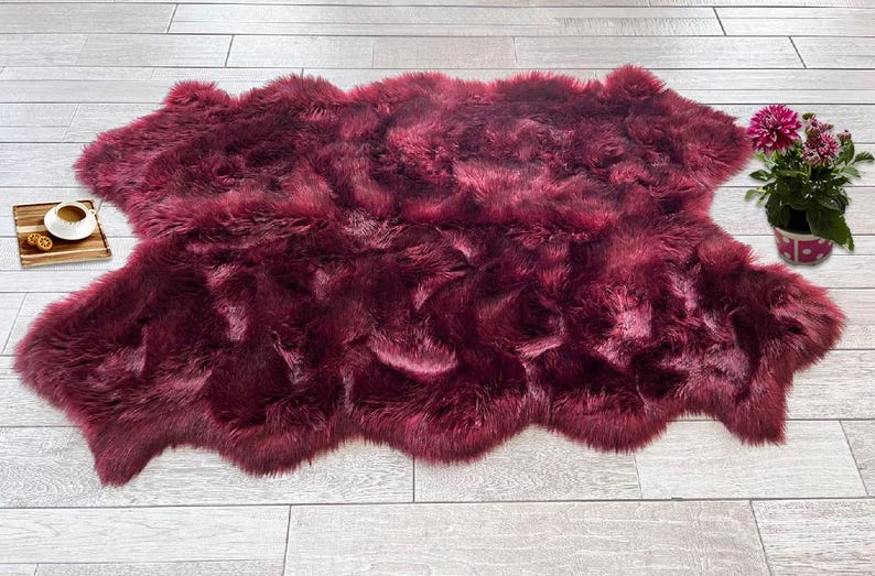 May include: A burgundy-colored sheepskin rug with a soft, textured appearance. The rug is irregularly shaped and lies on a light-colored wooden floor. A small wooden tray with a cup and cookies sits to the left, and a potted plant with pink flowers is on the right.