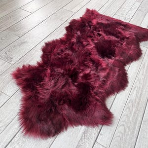 May include: A plush, burgundy-colored sheepskin rug. The rug has a soft, furry texture and an irregular shape. It is lying on a light gray wooden floor, creating a contrast in color and texture. The rug appears to be made of natural fibers.