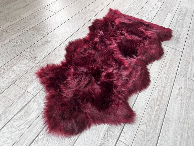 May include: A plush, burgundy-colored sheepskin rug. The rug has a soft, furry texture and an irregular shape. It is lying on a light gray wooden floor, creating a contrast in color and texture. The rug appears to be made of natural fibers.