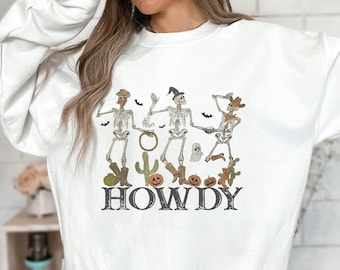May include: White sweatshirt featuring a design of three skeletons dressed as cowboys dancing with pumpkins, cacti, and the word "HOWDY" printed below.