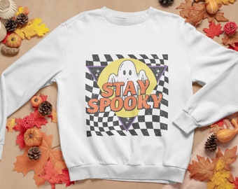 Może przedstawiać: Biała bluza z czarno-białym wzorem w kratkę, żółtym kołem z białym duchem i napisem "STAY SPOOKY" w pomarańczowych literach.