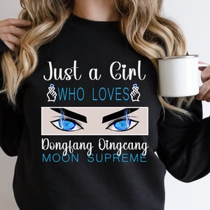 Könnte beinhalten: Schwarzer Pullover mit dem Text "Just a Girl Who Loves" mit einem Handgestensymbol auf jeder Seite des Textes. Unter dem Text befindet sich eine Grafik mit zwei blauen Augen mit Blitzen. Der Text "Dongfang Dingcang Moon Supreme" befindet sich unter den Augen.