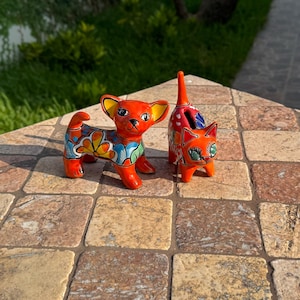May include: Two ceramic figurines: a dog and a cat. The dog is orange with yellow ears and a colorful floral design on its body. The cat is also orange with green eyes and a tall, slender tail. Both are on a stone surface.