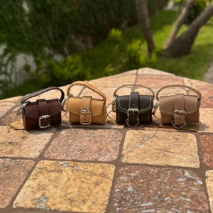 May include: Four miniature leather handbags in shades of brown, tan, and black are displayed. Each bag features a small handle, a buckle closure, and a metal ring. The bags are arranged on a textured, multi-colored surface.