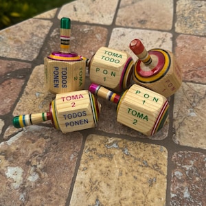 May include: A collection of wooden spinning tops with colorful stripes and text. The tops have a cylindrical body and a handle. The text on the sides reads "TOMA TODO", "PON 1", "TOMA 2", and "TODOS PONEN".