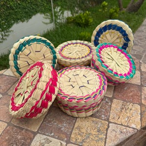 May include: A collection of woven baskets in various sizes and colors. The baskets are made of natural fibers and feature colorful accents in pink, green, blue, and yellow. The baskets are round and have lids.