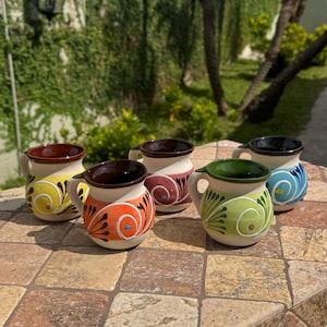 May include: Five handcrafted ceramic mugs with handles, each featuring a unique design. The mugs have a cream-colored base with colorful patterns in yellow, orange, red, green, and blue. The interiors are dark brown or black.