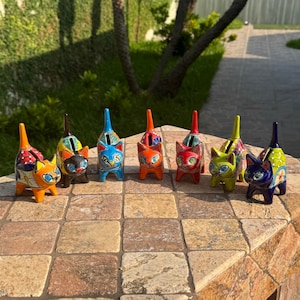 May include: A collection of colorful ceramic cat figurines. Each cat has a unique design, with vibrant colors like orange, blue, red, and yellow. The cats are standing on a tiled surface, showcasing their detailed craftsmanship.
