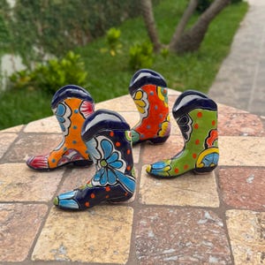 May include: Four colorful ceramic cowboy boots. Each boot features a unique design with vibrant colors like orange, blue, green, and red. The boots are decorated with floral and geometric patterns, and are displayed on a stone surface.