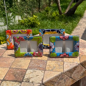 May include: Four colorful ceramic picture frames with floral designs. The frames are rectangular with a central opening for a photo. Each frame has a different color scheme, including orange, green, blue, and yellow, with intricate floral patterns.