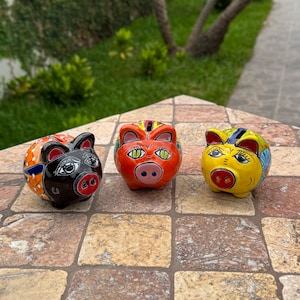 May include: Three colorful ceramic piggy banks. The banks are painted with vibrant colors and patterns, including orange, black, yellow, and blue. Each bank has a slot on top for coins and a cute pig face design.