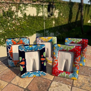 May include: A collection of five rectangular ceramic picture frames. Each frame features a vibrant floral design in various colors, including blue, orange, red, and yellow. The frames are arranged on a stone surface, showcasing their decorative patterns.