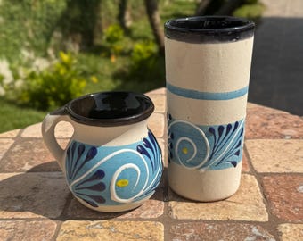 May include: Two handmade ceramic mugs. One is a small, handled mug with a black interior and a white exterior decorated with blue and yellow floral designs. The other is a tall, cylindrical mug with a black rim and blue accents.