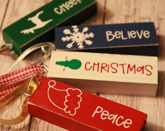 May include: Rectangular wooden Christmas ornaments in green, blue, white, and red. Each block has a festive word or image: "Cheer," a snowflake, "Believe," a green ornament, "Christmas," and "Peace." Ribbons are attached for hanging.
