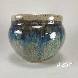 May include: A rounded ceramic bowl featuring a speckled beige rim that transitions into a deep blue and sage green glaze with vertical drip patterns. The base is a light tan color, and the text # 2571 is visible in the bottom right corner.